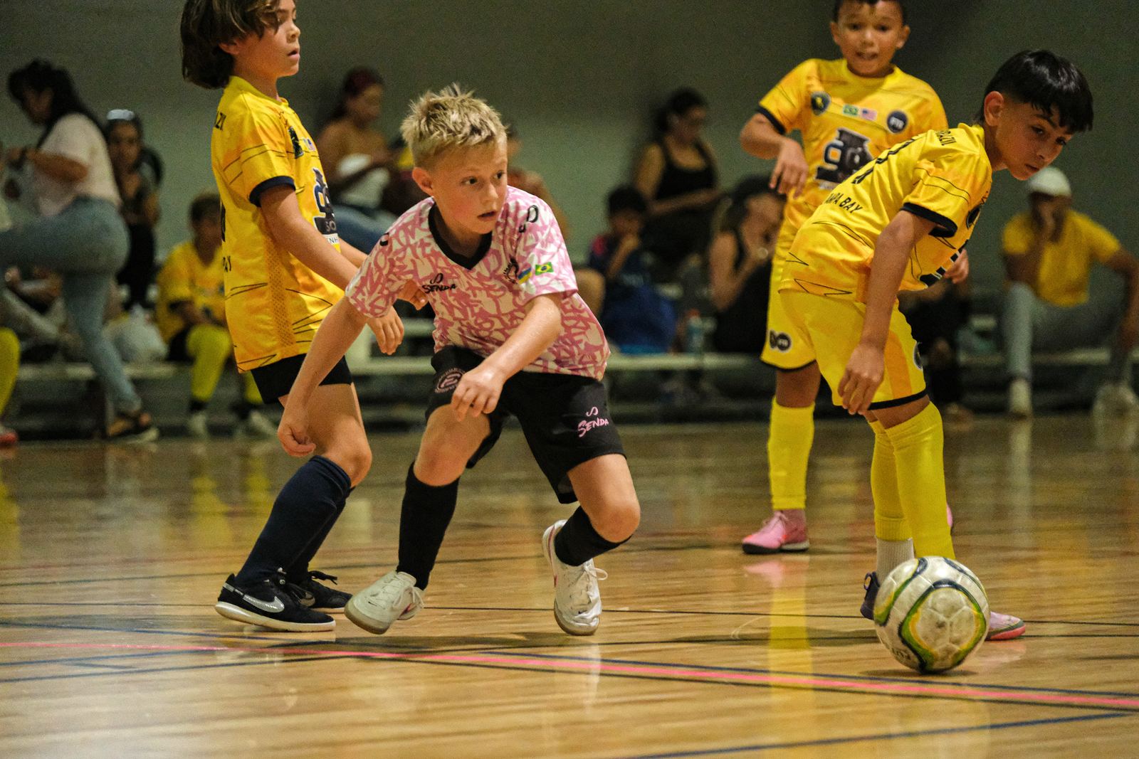 Youth indoor soccer players chasing the ball in front of a packed bleacher crowd